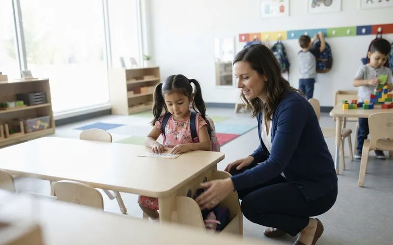 Child with a small backpack walking hand in hand with a parent toward a kindergarten classroom entrance